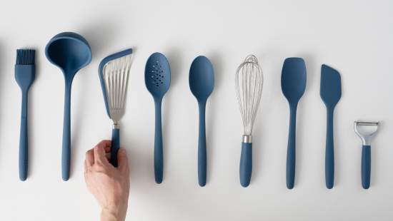 Row of blue silicone-handled kitchen utensils on white background; hand lifting a stainless slotted turner