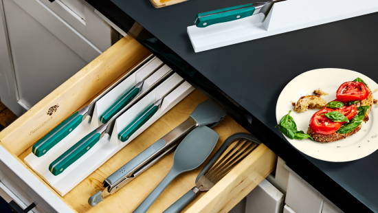 Bamboo drawer with teal-handled knives in white slots, gray utensils, black countertop, plate of tomato and basil toast.