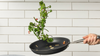 A Misen Carbon Nonstick™ frying pan, held by a hand on the right, is shown mid-toss, with broccolini and some sliced chili peppers and garlic cloves suspended in the air above the pan. The background is a white subway tile wall.