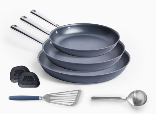 This image depicts the Misen Carbon Nonstick™ Complete Set. It features three Carbon Nonstick™ Frying Pans of varying sizes, a blue-handled stainless steel slotted fish spatula, a measuring chef spoon and two pan scrapers. The pans and utensils are arranged on a white background.