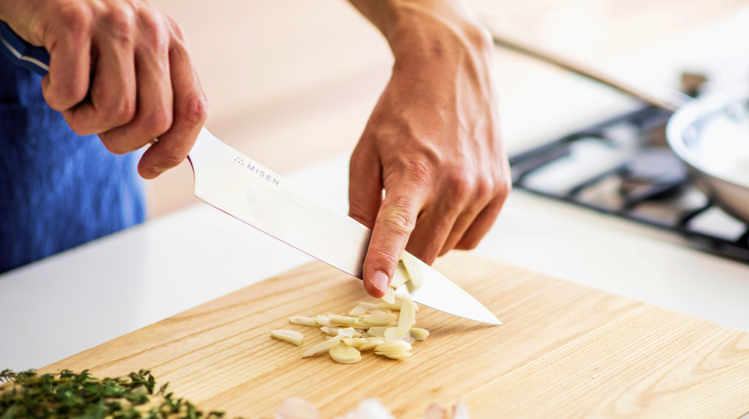 Hands using a Misen chef's knife to slice garlic on a wooden cutting board near a stovetop.
