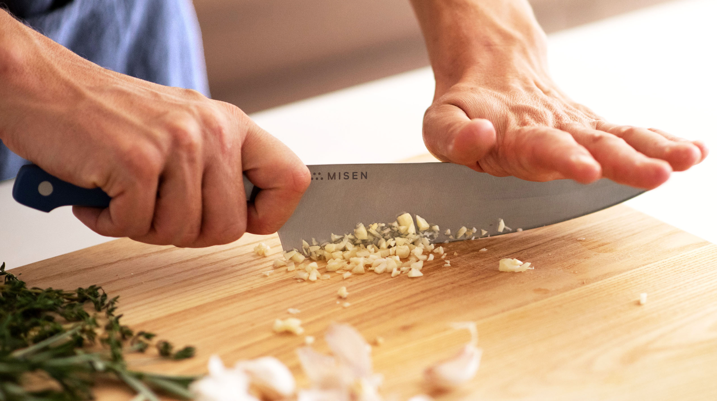 Hands using a Misen chef's knife to mince garlic on a wooden cutting board with herb sprigs nearby