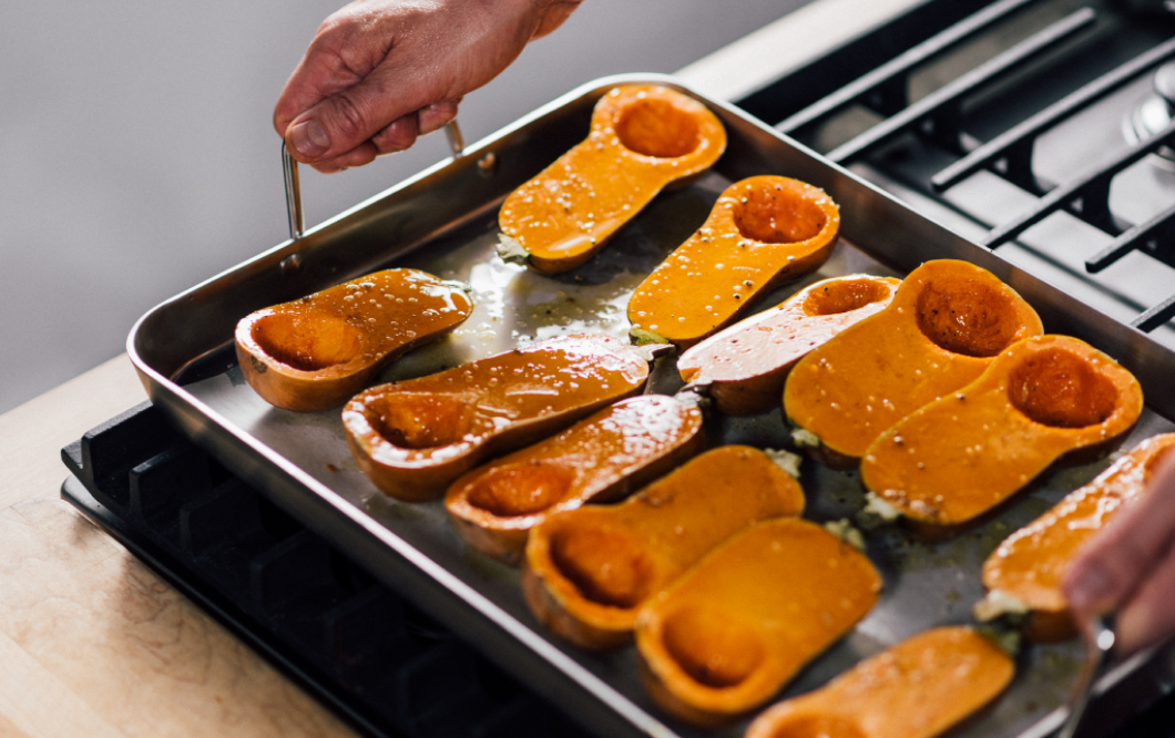 Hand holding a metal roasting pan filled with halved butternut squash cavities facing up on a stovetop