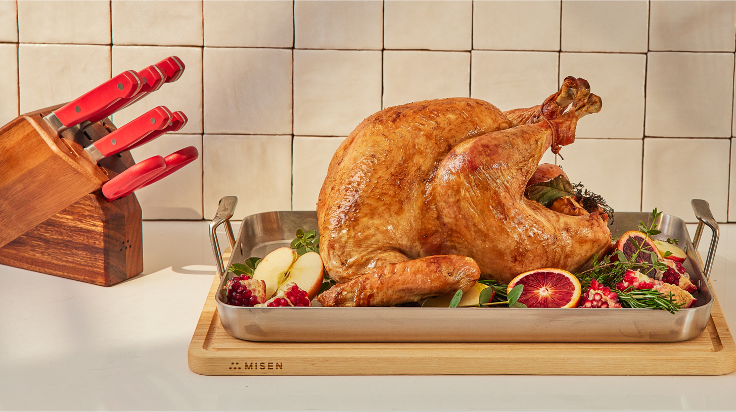 Roasted turkey on stainless pan on wooden board labeled MISEN, with fruit and herbs; red-handled knives at left.