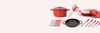 Red lidded Dutch oven, red shallow sauté pan, red-handled knife set, grill pan, and kitchen shears on pale background