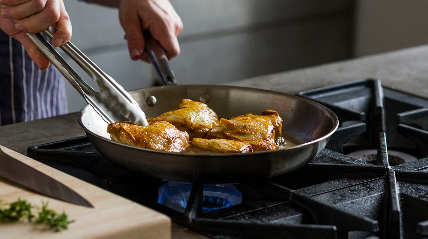 Hands using metal tongs to turn browned chicken thighs in a stainless steel skillet over a blue gas flame.