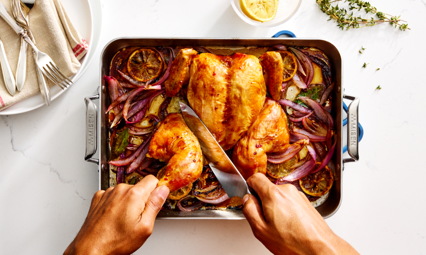 A roasted chicken in a Misen roasting pan surrounded by roasted red onions, lemon slices, and herbs. A person's hands are visible, using a knife to carve a leg and thigh from the chicken.