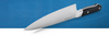 A close-up shot of a Misen 8 inch Chef's Knife with a black handle and silver ACUTO440 steel blade resting on a blueprint. The Misen logo is visible on the blade, and a faint outline of a knife is visible on the blueprint behind it.