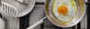 A close-up view of a Misen stainless steel frying pan with a single, perfectly fried egg (sunny-side up with pepper) resting on a black gas stovetop grate.