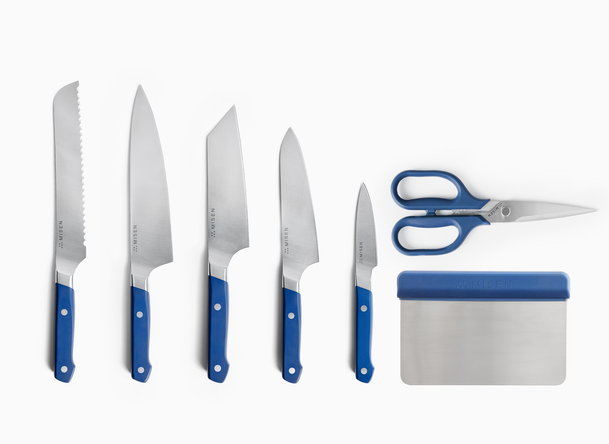 A high-angle shot on a white background shows five Misen ACUTO440 steel knives with blue handles, a pair of standard blue-handled Misen kitchen shears, and a Misen bench scraper. The knives are arranged from left to right in descending order of size: an 8 inch serrated knife, an 8 inch chef's knife, a 6 inch bunka knife, a 5.5 inch utility knife, and a 3.5 inch paring knife. To the right of the knives, the kitchen shears and bench scraper are stacked vertically.