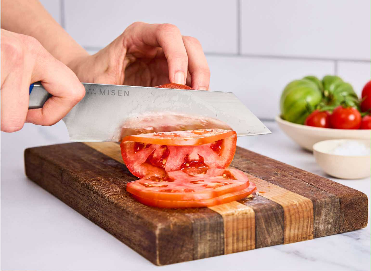 A person is shown slicing a red tomato on a wooden cutting board with a Misen 6 inch ACUTO440 steel Bunka knife. In the background, a bowl of green and red tomatoes, along with a small white bowl of salt, is visible.
