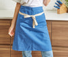 A blue Misen apron is worn on a chef and folded over to create a bistro apron. The chef is visible from the chest down and is leaning against a kitchen counter.