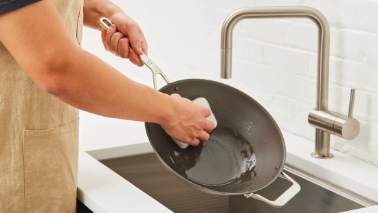 Person wearing beige apron scrubbing a dark nonstick pan with a white sponge over a stainless sink and faucet