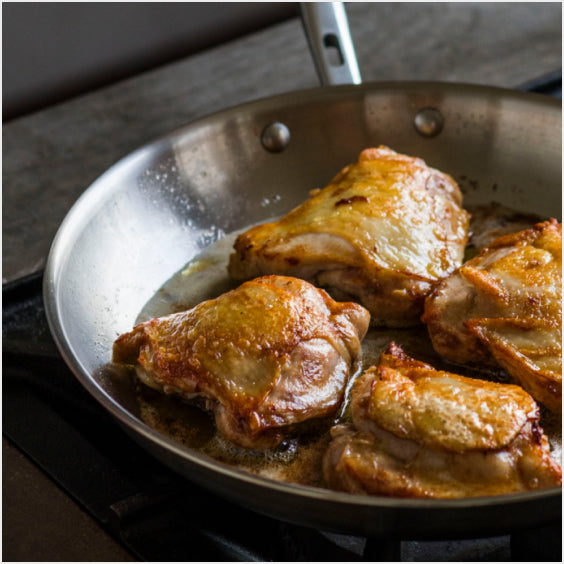 A close-up image of a misen stainless steel skillet on a stovetop containing three golden-brown chicken thighs sizzling in butter or oil.
