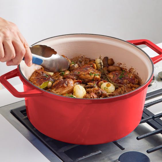 A hand using tongs to serve chicken pieces from a red Dutch misen oven on a stovetop. The dish contains chicken, mushrooms, pearl onions, and herbs, with a rich sauce.