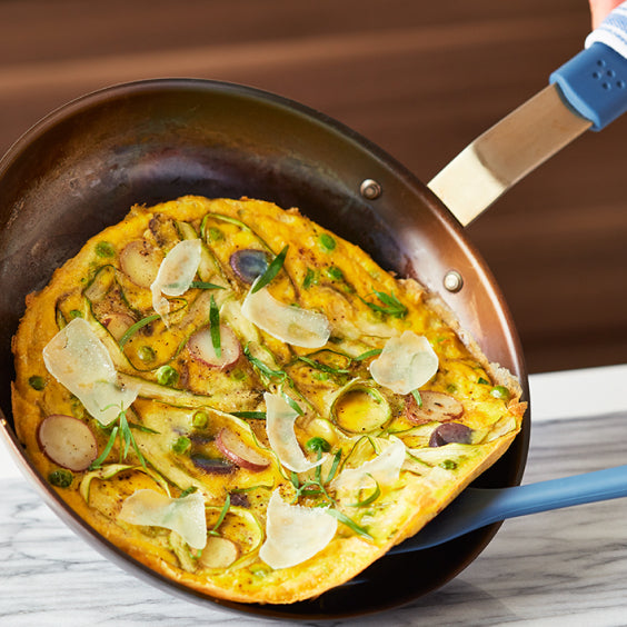 A copper misen skillet holds a colorful frittata garnished with shaved Parmesan, peas, radish slices, and fresh herbs. A blue-handled spatula is lifting a portion of the frittata, with a marble countertop in the background.