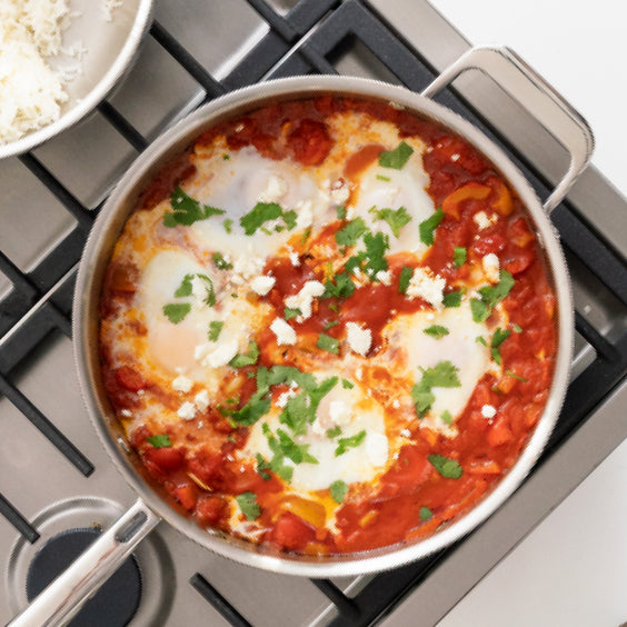 A misen skillet on a stovetop contains a dish of shakshuka with poached eggs, a rich tomato sauce, and crumbled feta cheese, garnished with fresh cilantro. A bowl of white rice is partially visible in the background.