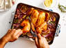 An overhead shot of a person carving a roasted chicken in a Misen Stainless Steel Roasting Pan. The chicken is surrounded by roasted red onions and lemon slices. The roasting pan has the 