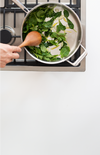 Hand stirring leafy greens and sliced garlic in stainless-steel pan on gas stovetop with wooden spoon