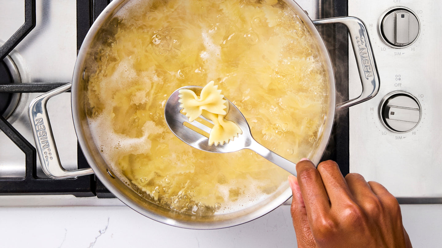 Bowtie farfalle pasta being tested for doneness in boiling water using a slotted spoon in a Misen stainless steel pan