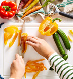 Hands slicing yellow bell pepper on white cutting board with fresh vegetables
