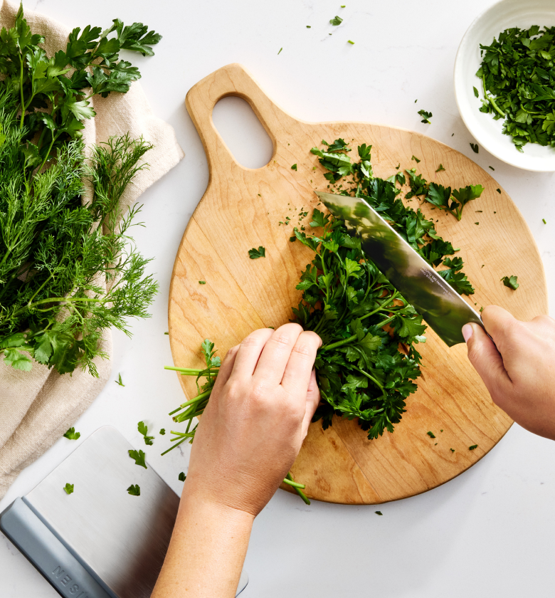 Hands chopping fresh parsley on a round wooden cutting board with dill and herbs nearby