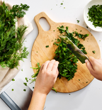 Hands chopping fresh parsley on a round wooden cutting board with dill and herbs nearby