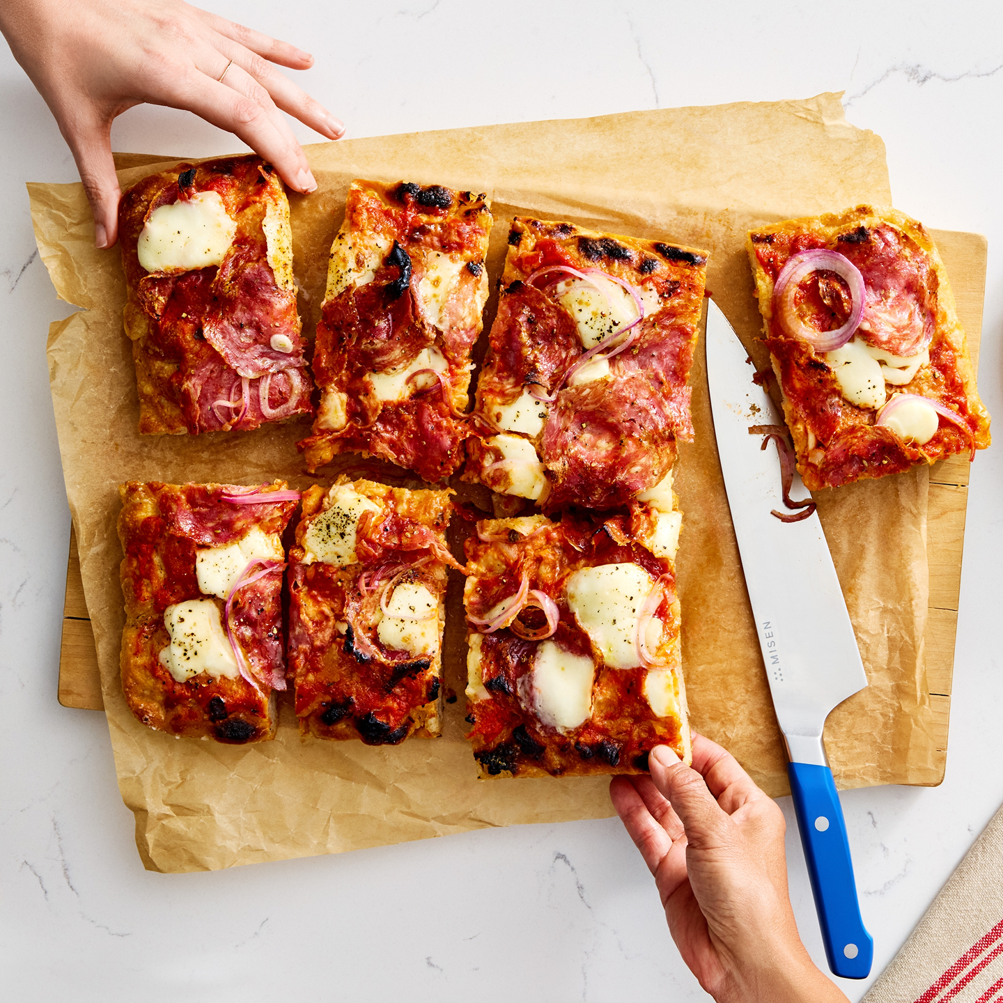 Sliced salami and mozzarella sheet pan pizza on parchment paper being cut with a Misen chef's knife