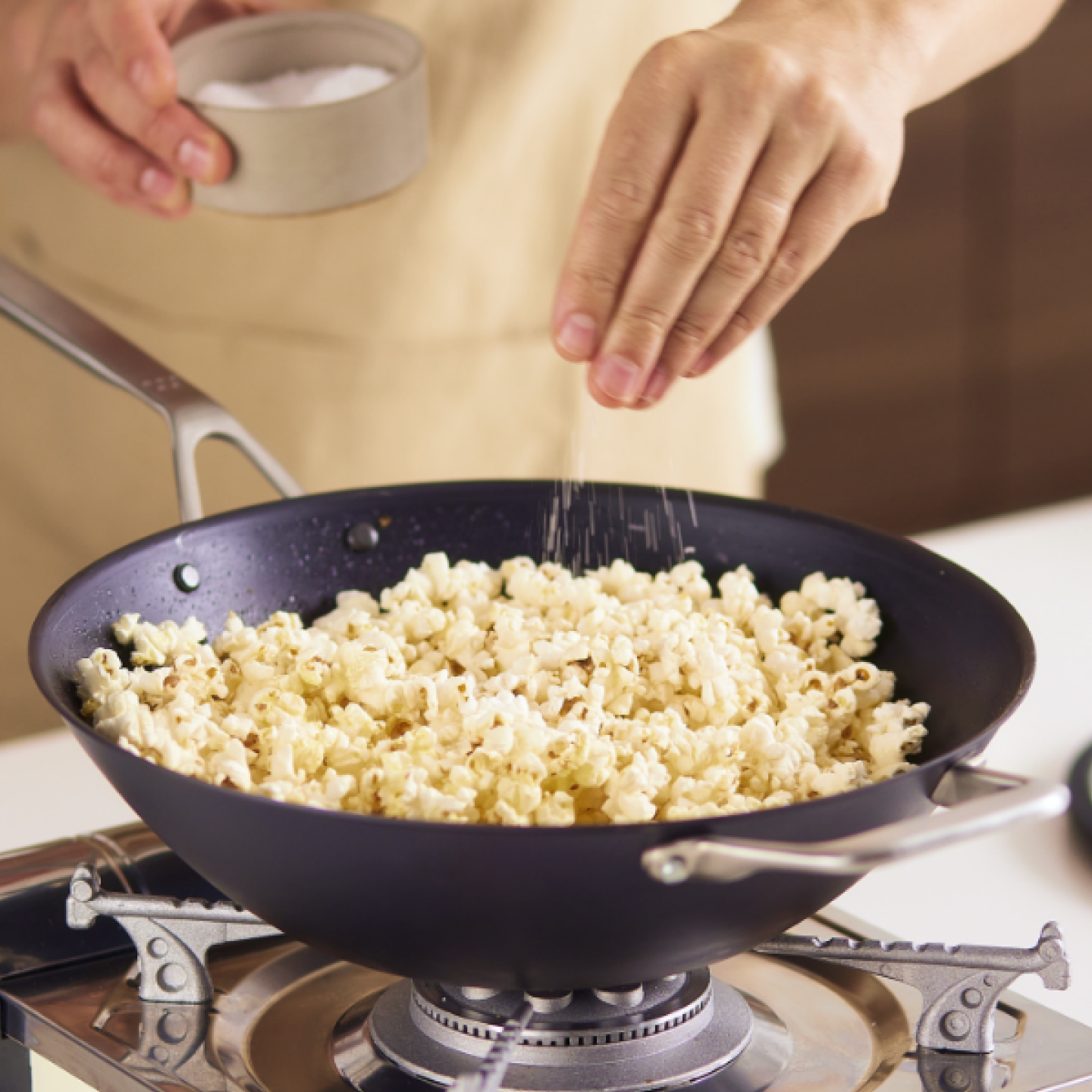 Person seasoning freshly popped popcorn with salt in a wok on gas stovetop