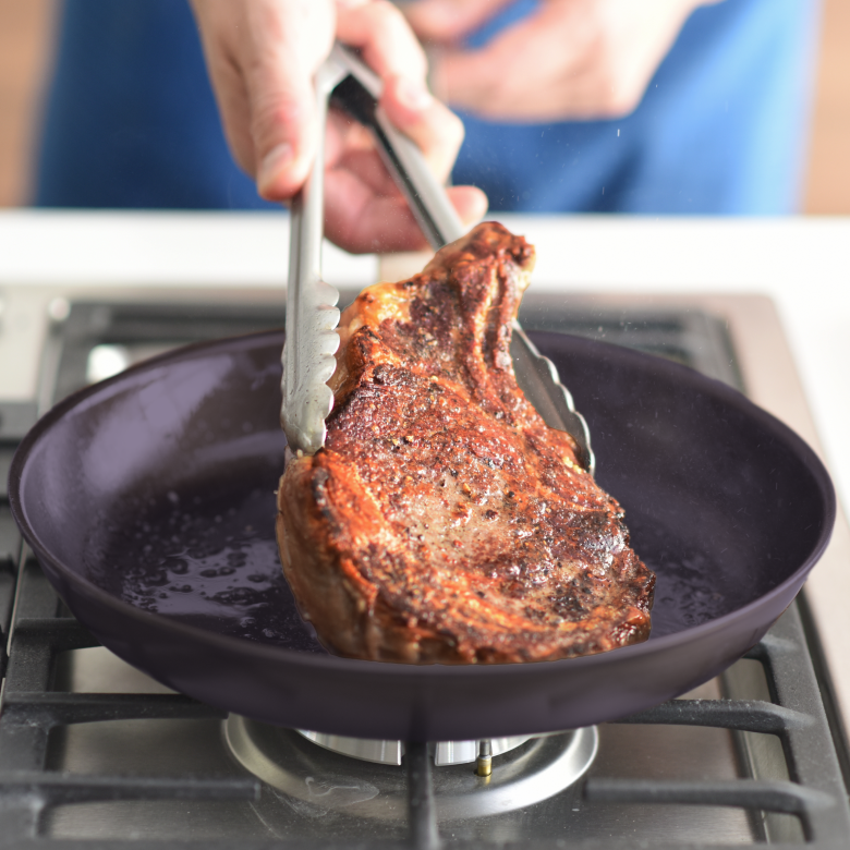 Searing a seasoned ribeye steak in a skillet on a gas stovetop with tongs