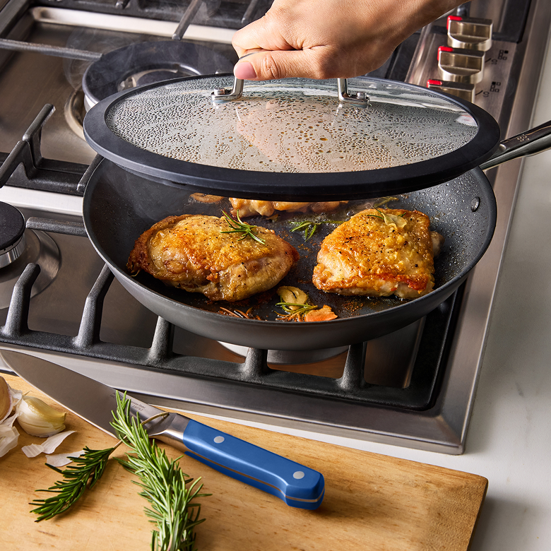 Golden seared chicken thighs cooking in nonstick skillet with glass lid on gas stove