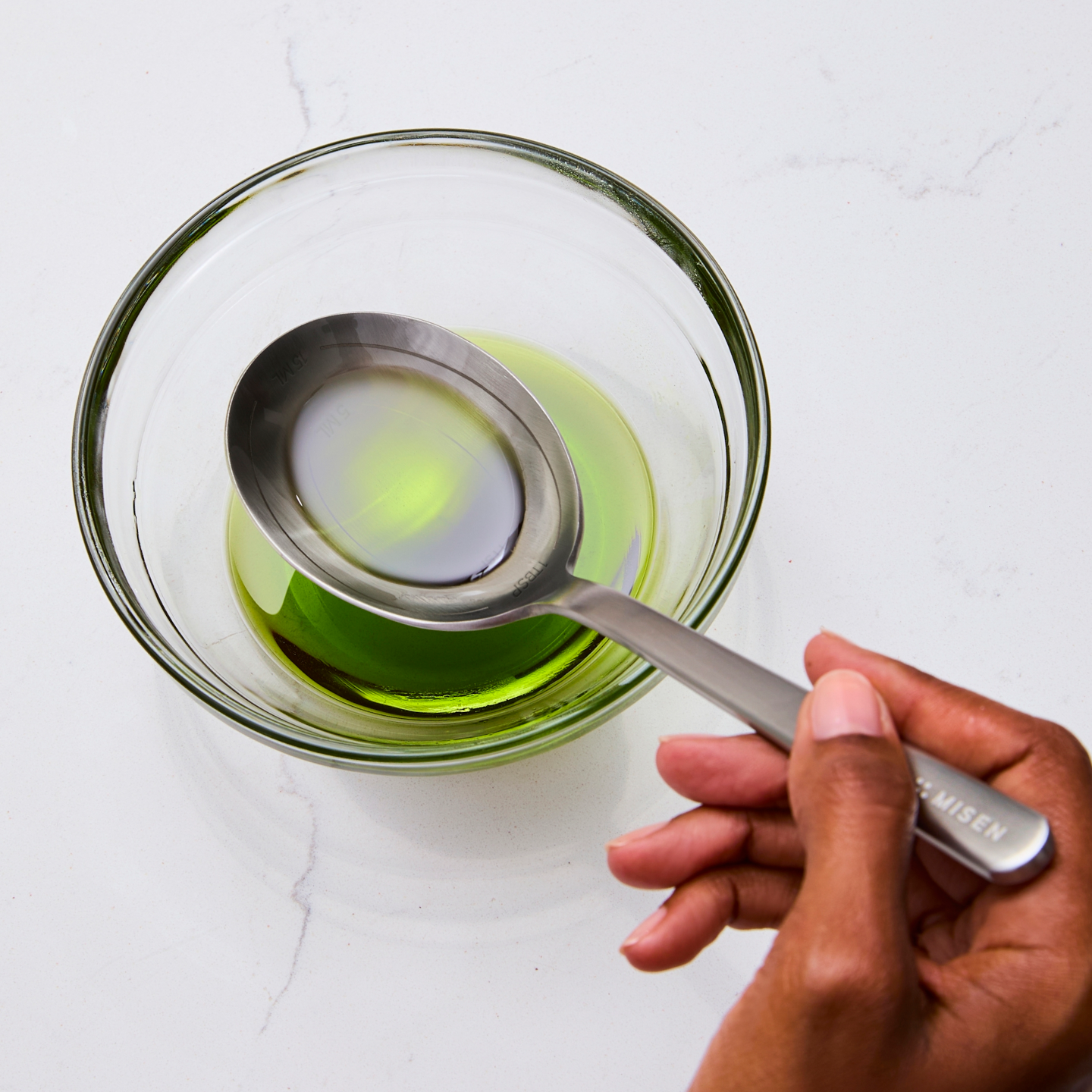 Measuring spoon adding oil to glass bowl with green liquid
