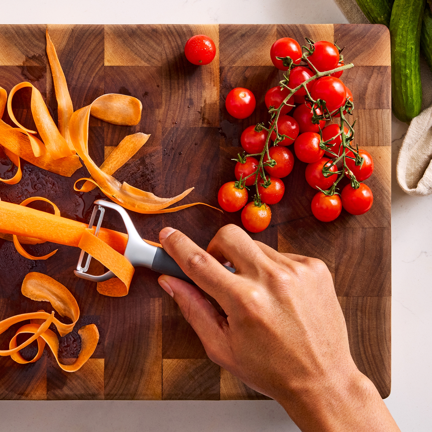 Hand peeling a carrot with a Y-peeler on a wood cutting board beside fresh cherry tomatoes