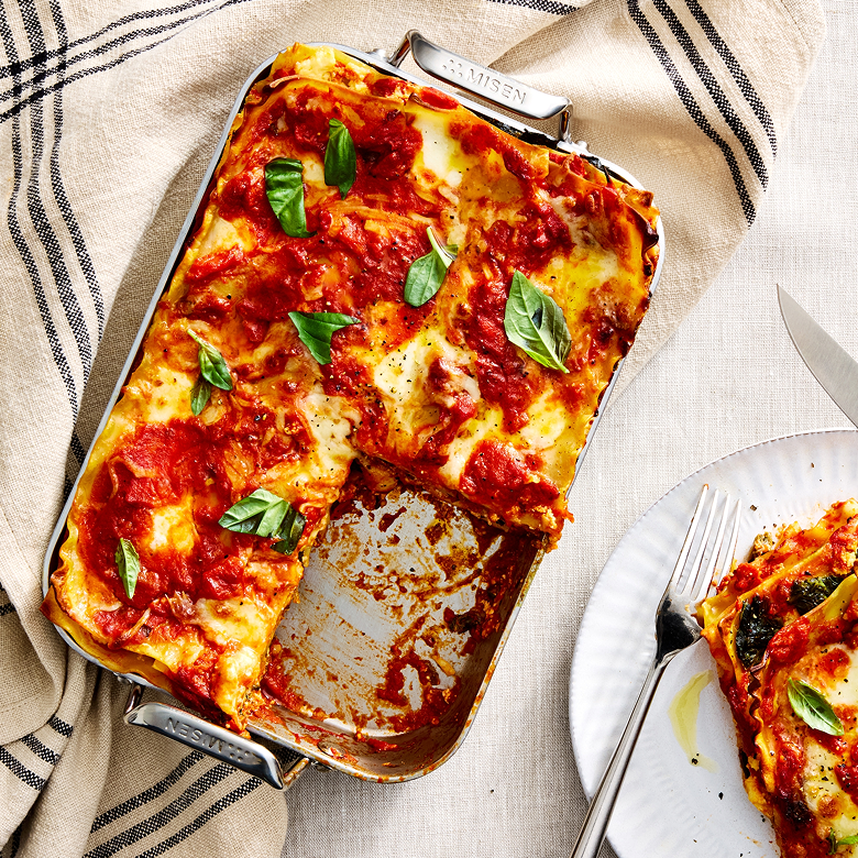 Homemade lasagna in a Misen stainless steel baking pan topped with tomato sauce, melted mozzarella, and fresh basil leaves, with a served slice on a plate