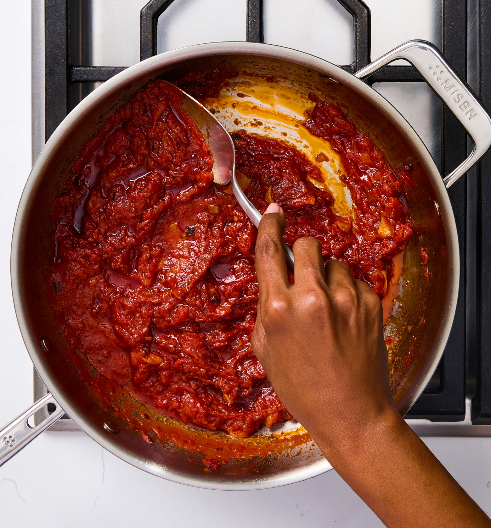 Hand stirring rich tomato sauce in a stainless steel Misen pan on gas stovetop