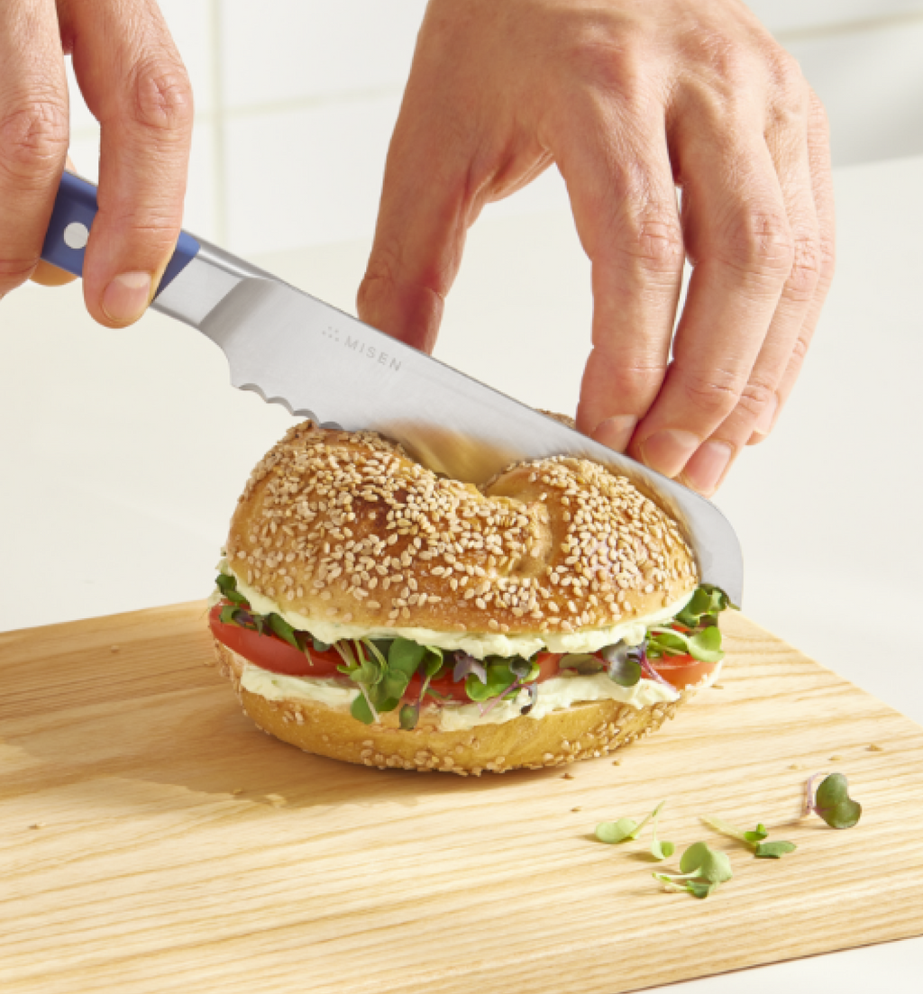 Misen serrated bread knife cutting a sesame bagel sandwich with cream cheese, tomato, and microgreens on a wooden cutting board