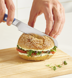 Misen serrated bread knife cutting a sesame bagel sandwich with cream cheese, tomato, and microgreens on a wooden cutting board