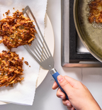 Crispy potato latkes being transferred from frying pan to paper towel-lined plate with slotted spatula