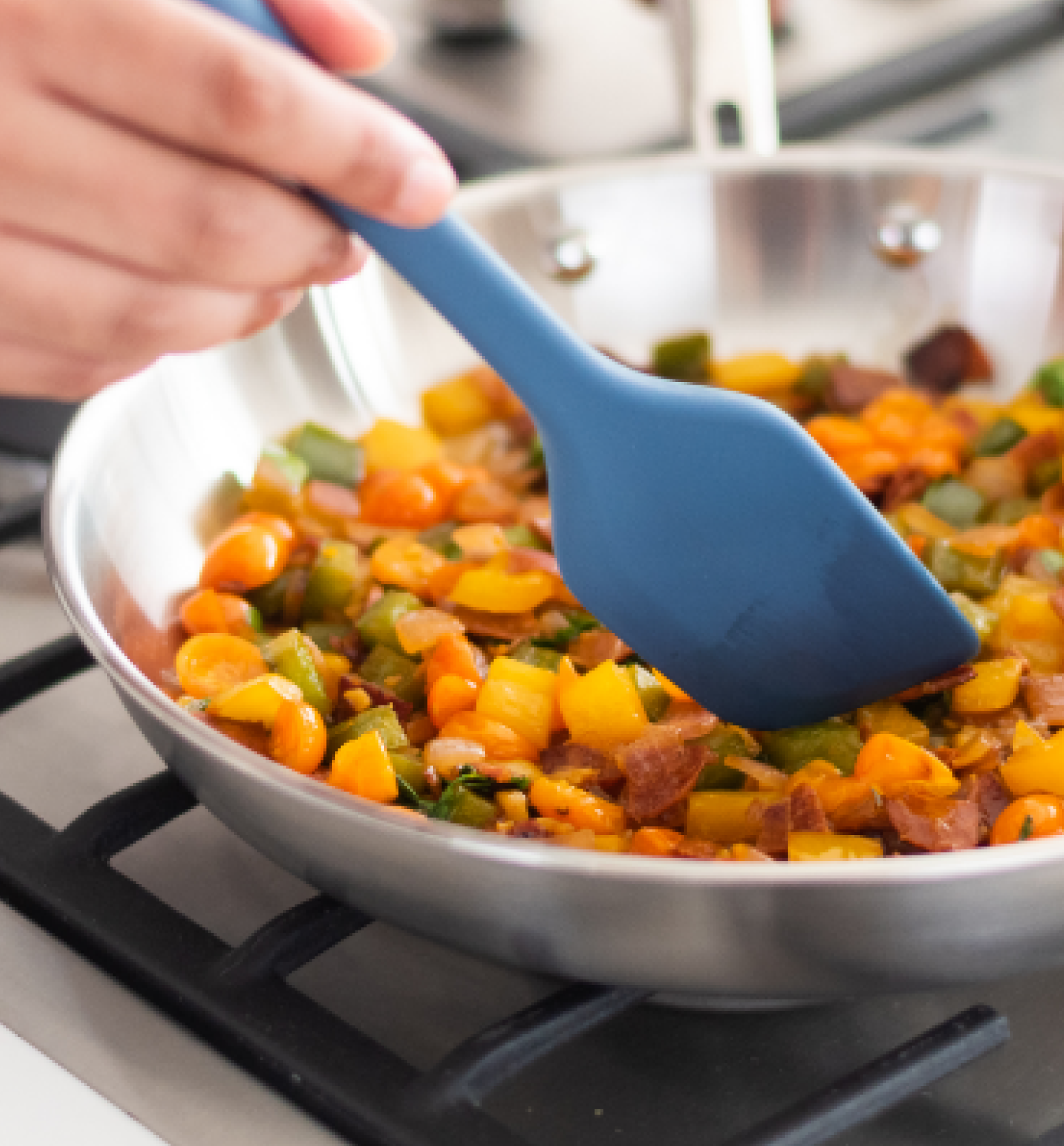 Sautéing colorful vegetables and bacon in a stainless steel skillet with a blue silicone spatula