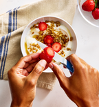 Hands slicing fresh strawberries over a yogurt granola bowl
