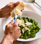 Hands shaving Parmesan cheese over green pesto rigatoni pasta in white bowl