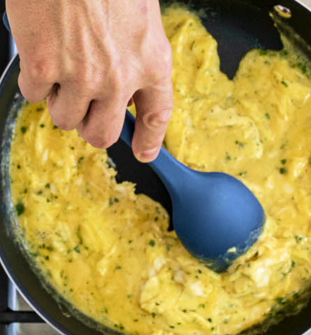 Scrambled eggs with herbs being stirred in nonstick pan with blue silicone spoon