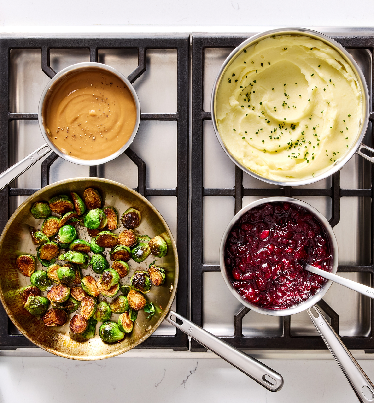 Thanksgiving side dishes on stovetop including mashed potatoes, roasted Brussels sprouts, cranberry sauce, and gravy