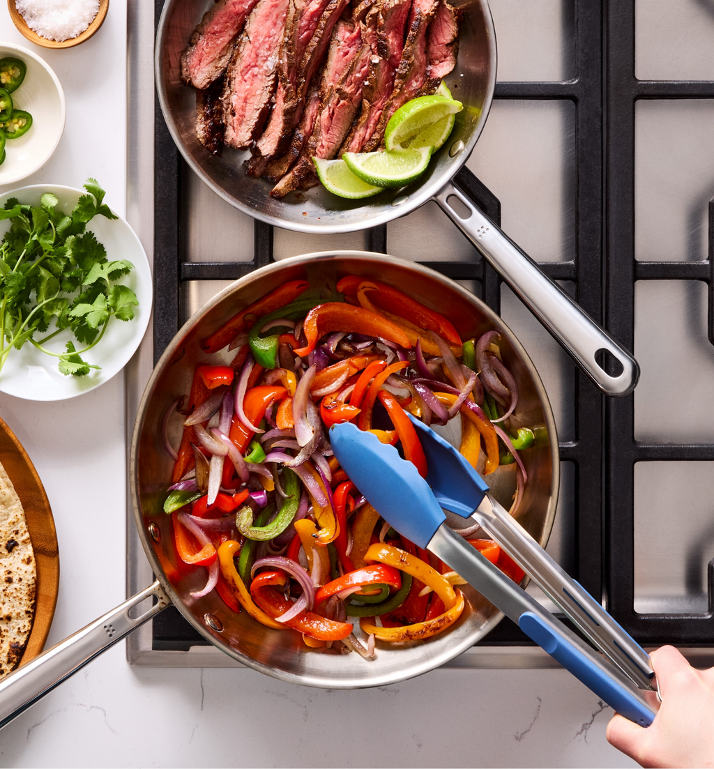Steak fajitas with sautéed peppers and onions cooking in stainless steel pans on gas stovetop