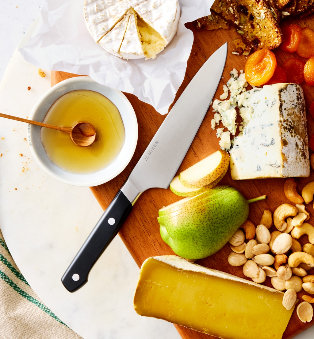 Misen chef's knife on wooden charcuterie board with brie, blue cheese, pear, nuts, honey, and dried apricots