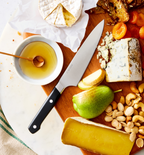Misen chef's knife on wooden charcuterie board with brie, blue cheese, pear, nuts, honey, and dried apricots