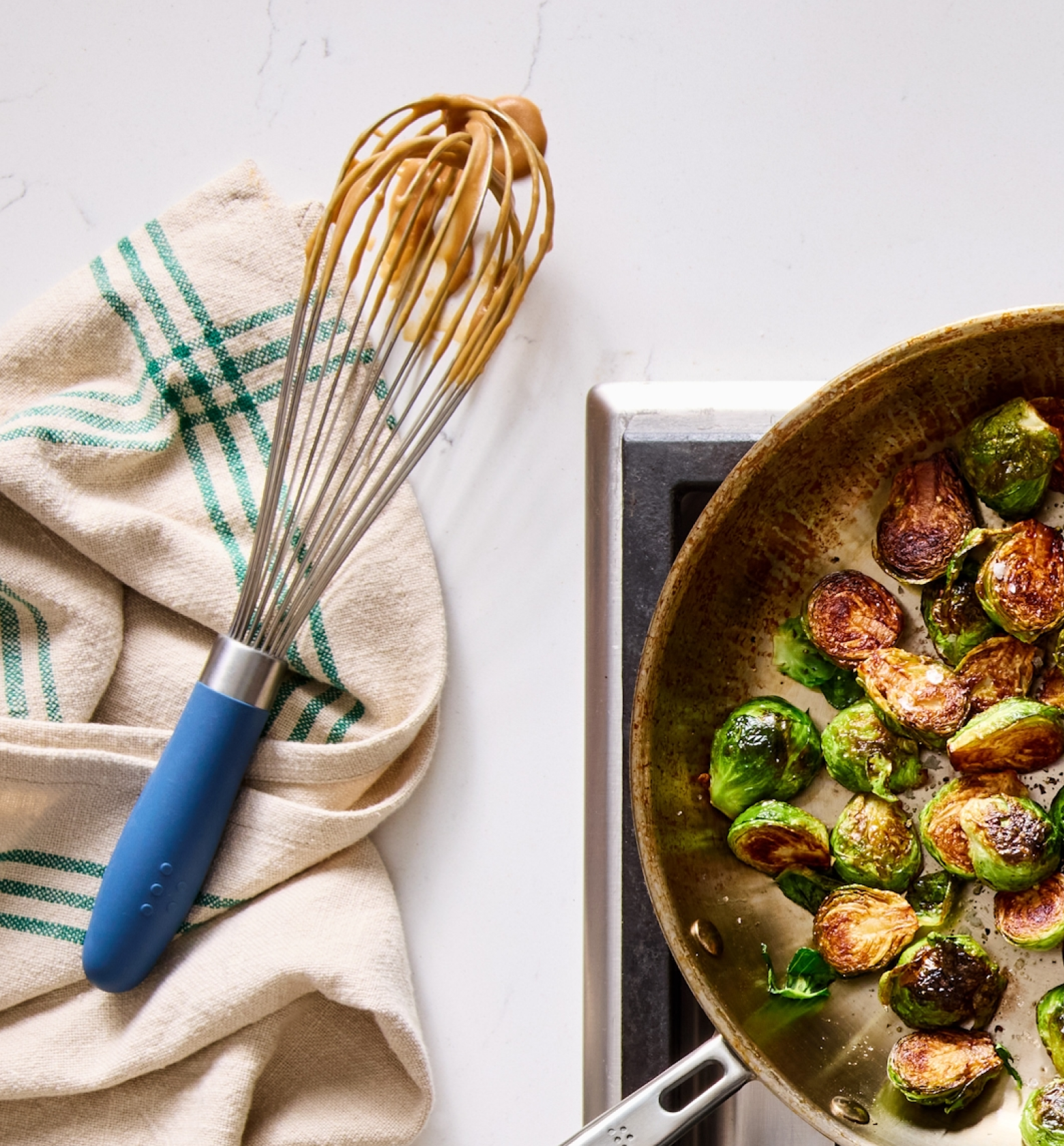 Sautéed Brussels sprouts in a skillet on stovetop with a blue-handled whisk and kitchen towel