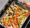 Roasting pan with halved baby potatoes, carrot sticks, parsnip pieces, garlic cloves and sprigs of thyme; hands on handles