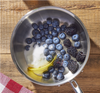 Stainless steel saucepan with blueberries, blackberries, granulated sugar and a lemon peel on wooden table.
