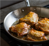 Four browned chicken thighs searing in a stainless steel pan on a gas stove.